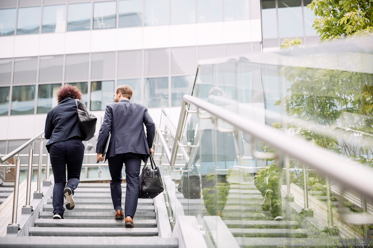 Two professionals in business attire ascending an outdoor staircase with glass railings, set against a modern building with large windows and greenery, representing a corporate or urban work environment.