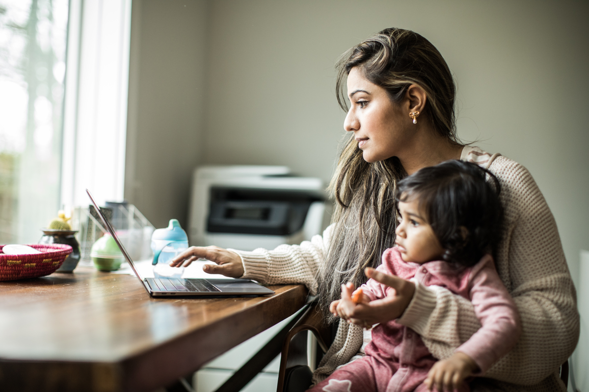 Person working on a laptop at a wooden table while holding a young child. The table is scattered with everyday items like a fruit bowl, baby bottle, and papers, set in a naturally lit home environment.