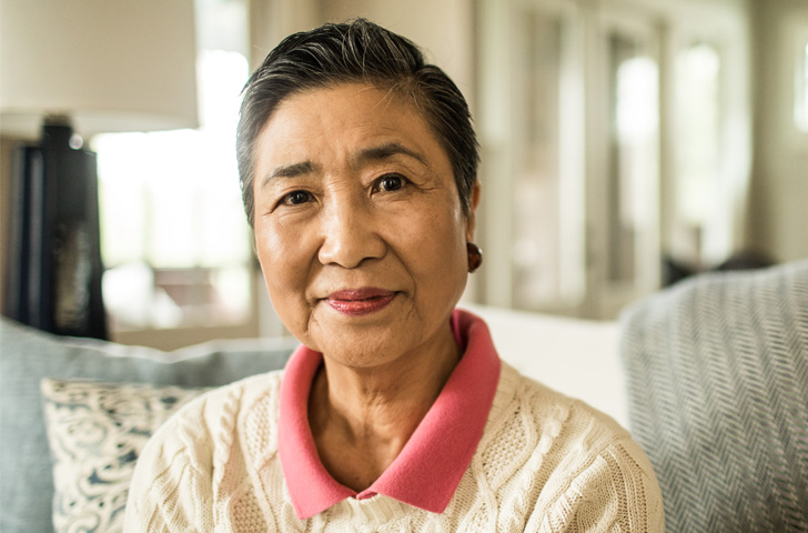 Older woman sitting indoors. She is wearing a cream colored cable knit sweater and a pink collared shirt