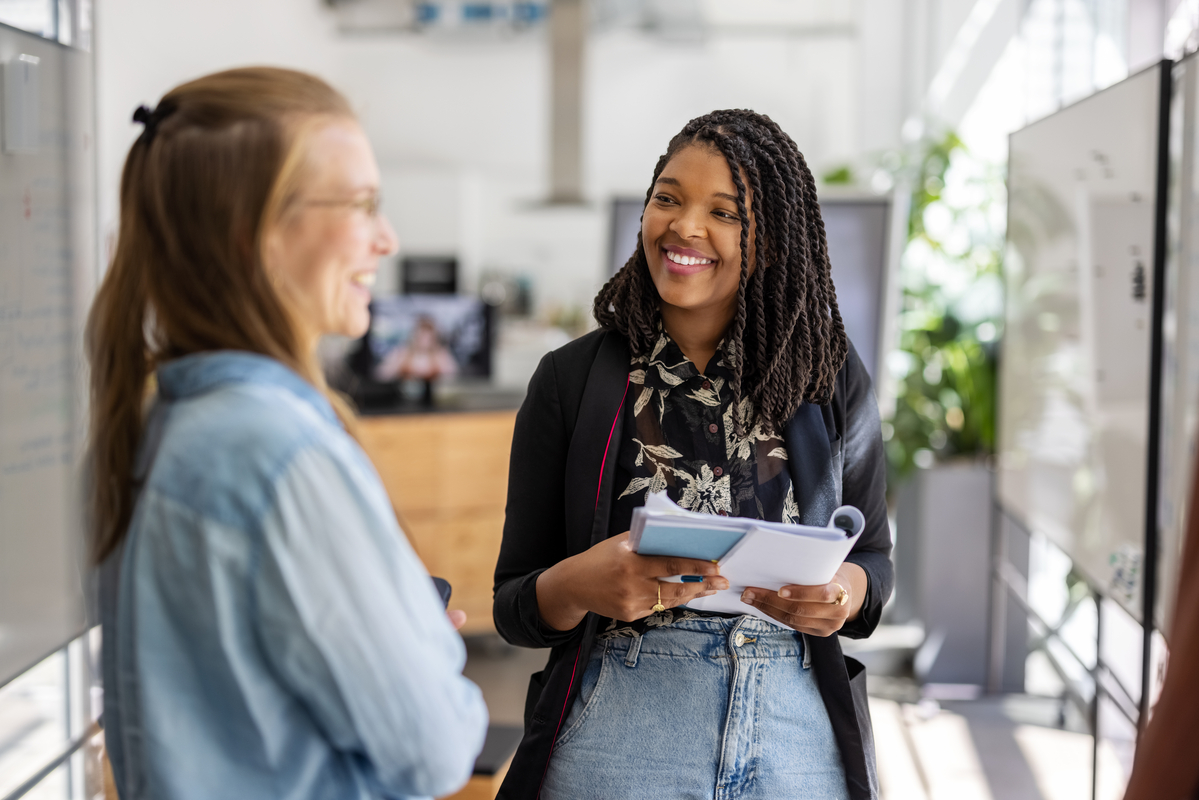 : Two female colleagues discussing new business plan in office. Startup businesswoman talking with trainee employee holding a book in hybrid open plan office.