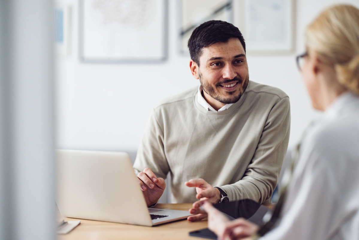  Caucasian businessman is sitting in an office and having a meeting with a female colleague. He might be working from home. It's morning or later in the day. He is smiling. A laptop is in front of him and the woman is holding a digital tablet.