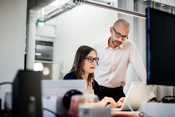 businesswoman is working late on her notebook sitting at a desk in a modern office room, while a colleague is standing next to her, discussing something. There's more office equipment on the desk.