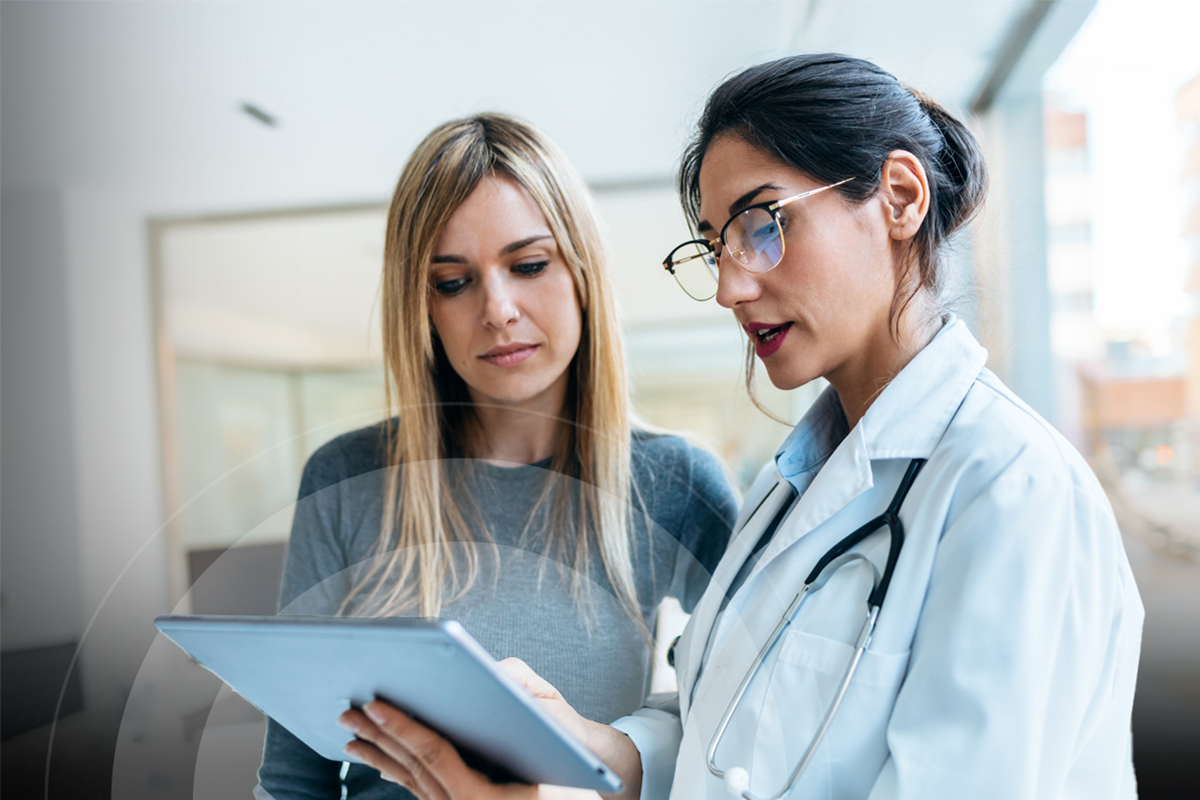 Provider talking to a patient in an exam room. The provider has a tablet in her hand while pointing at data